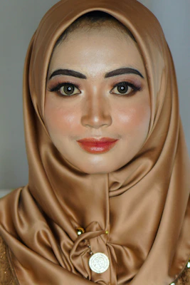 A portrait of a person wearing a silk brown hijab with elaborate makeup, including bold eyeliner and red lipstick. A decorative pendant is visible on the hijab.