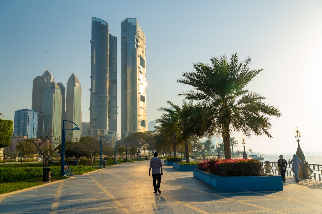 A lone pedestrian walks past high-rise towers on the Abu Dhabi Corniche at sunset.