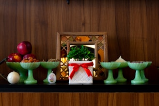 A display of decorative items on a wooden shelf includes a potted plant in a white container with a red ribbon, several green pedestal bowls holding various items like red apples and nuts, and a small, round candle. A framed decorative mirror in the background adds to the arrangement.