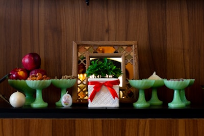 A display of decorative items on a wooden shelf includes a potted plant in a white container with a red ribbon, several green pedestal bowls holding various items like red apples and nuts, and a small, round candle. A framed decorative mirror in the background adds to the arrangement.
