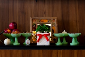 A display of decorative items on a wooden shelf includes a potted plant in a white container with a red ribbon, several green pedestal bowls holding various items like red apples and nuts, and a small, round candle. A framed decorative mirror in the background adds to the arrangement.