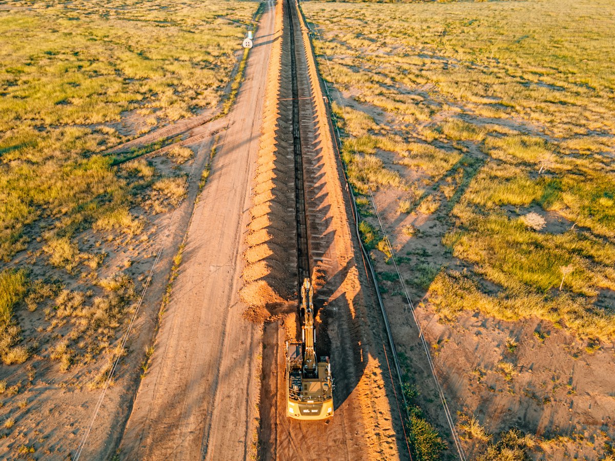 Aerial view of an excavator working along a straight earthwork trench with soil stockpiled beside it.