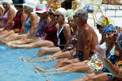 A group of elderly individuals, both men and women, are sitting side by side along the edge of a swimming pool with their feet in the water. They are wearing vibrant, colorful swimsuits and sun hats. The mood appears relaxed and social, with some people smiling and others in conversation.