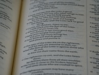 A close-up of an open book displaying text in a serif font. The text appears to be a passage from a religious or philosophical text, with numbered verses and bolded divine name references. The focus is on one page, with the other page slightly visible in the background.