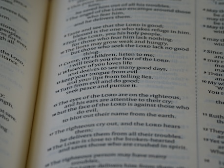 A close-up of an open book displaying text in a serif font. The text appears to be a passage from a religious or philosophical text, with numbered verses and bolded divine name references. The focus is on one page, with the other page slightly visible in the background.