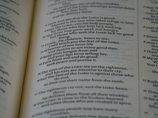 A close-up of an open book displaying text in a serif font. The text appears to be a passage from a religious or philosophical text, with numbered verses and bolded divine name references. The focus is on one page, with the other page slightly visible in the background.