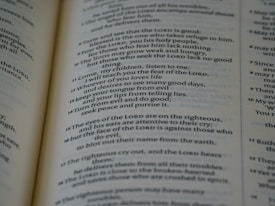 A close-up of an open book displaying text in a serif font. The text appears to be a passage from a religious or philosophical text, with numbered verses and bolded divine name references. The focus is on one page, with the other page slightly visible in the background.