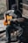 A person wearing a mask and a beanie plays an acoustic guitar while seated on a wooden stool. The setting appears rustic, with wooden walls and rough stone flooring, creating a cozy and intimate atmosphere. Natural light filters in, highlighting the person's hands and guitar.