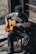 A person wearing a mask and a beanie plays an acoustic guitar while seated on a wooden stool. The setting appears rustic, with wooden walls and rough stone flooring, creating a cozy and intimate atmosphere. Natural light filters in, highlighting the person's hands and guitar.