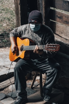 A person wearing a mask and a beanie plays an acoustic guitar while seated on a wooden stool. The setting appears rustic, with wooden walls and rough stone flooring, creating a cozy and intimate atmosphere. Natural light filters in, highlighting the person's hands and guitar.