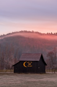 A serene landscape features a rustic barn with the phrase 'Love you to the moon and back' painted on its side, set against a backdrop of softly illuminated hills at sunset. Mist lingers over the forested hills, adding a mystical touch to the scene.
