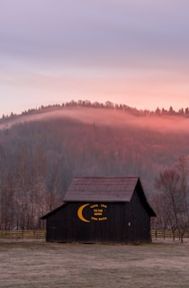 A serene landscape features a rustic barn with the phrase 'Love you to the moon and back' painted on its side, set against a backdrop of softly illuminated hills at sunset. Mist lingers over the forested hills, adding a mystical touch to the scene.