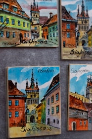 A collection of colorful painted artworks depicting a picturesque street scene with tall, ornate buildings. The images highlight a tower surrounded by historic architecture in bright shades of orange, yellow, blue, and green. The word 'Sighisoara' and 'Romania' are prominently displayed.