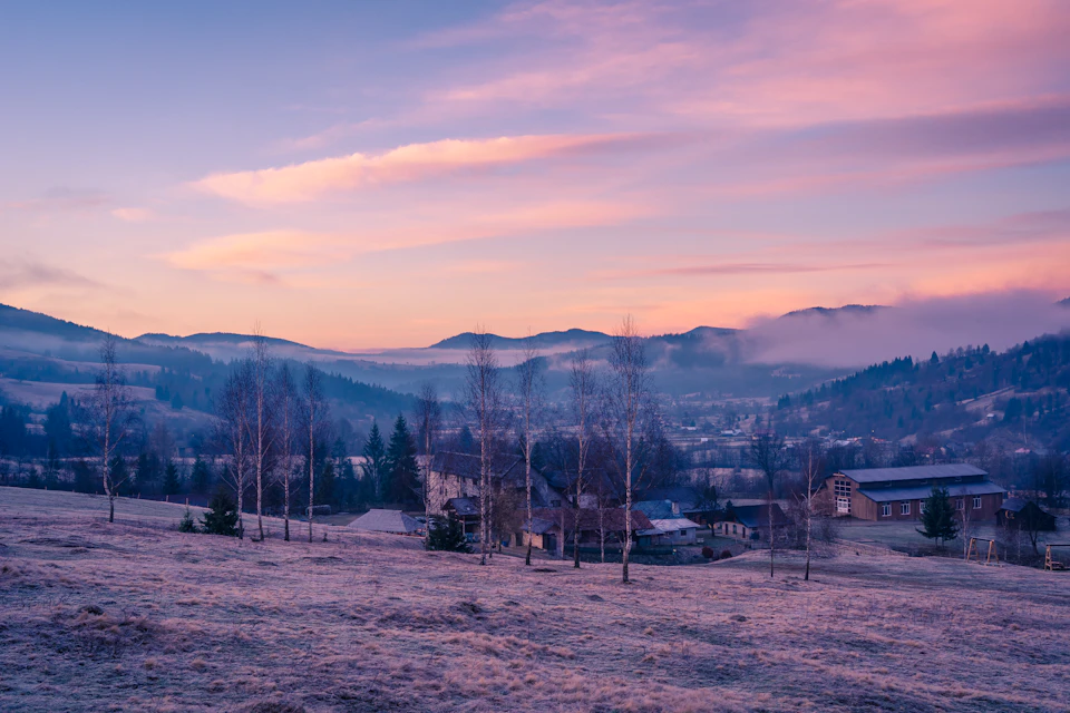 The image depicts a serene winter landscape with a small town nestled amidst snow-covered mountains and fields. The sky is painted in hues of pink, indicating either sunrise or sunset. The town features houses and buildings scattered across the landscape, their rooftops blanketed by snow. The trees surrounding the town are bare, suggesting that it's late autumn or winter. The image captures a tranquil moment during this season, with the sun casting its warm glow over the snowy terrain.

The perspective of the photo is from ground level, looking up at the town and mountains in the background. This viewpoint provides a sense of scale and grandeur to the scene, emphasizing the vastness of the landscape and the smallness of the town within it. The colors in the image are predominantly white, gray, and pink, reflecting the cold winter weather and the warm glow of the setting sun or sunrise.