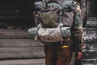 A person wearing a camouflage jacket and carrying a large backpack. The backpack has a rolled-up sleeping mat and a cloth with a hand-drawn peace sign and the words 'for peace' on it. The image suggests themes of travel or protest.