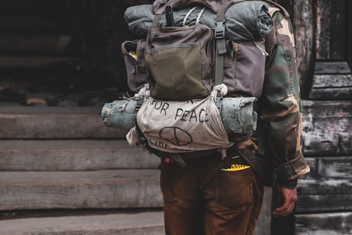 A person wearing a camouflage jacket and carrying a large backpack. The backpack has a rolled-up sleeping mat and a cloth with a hand-drawn peace sign and the words 'for peace' on it. The image suggests themes of travel or protest.