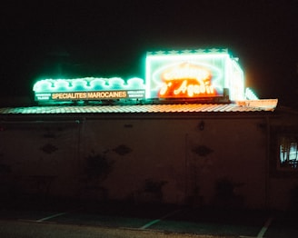 Neon signs in vibrant green and red illuminate the exterior of a Moroccan restaurant at night. The sign advertises specialties like couscous and tagines.