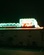 A dimly lit building with a brightly illuminated neon sign on the roof displaying text in French. The signs feature green and red hues, advertising Moroccan specialties. The atmosphere suggests a restaurant or caf&eacute; setting, with a dark, night-time background.