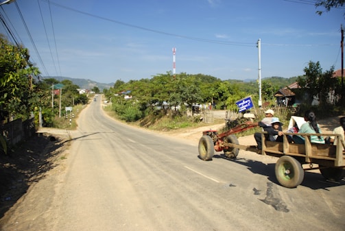 A rural road stretches forward with a tractor pulling a wooden cart on the right side. The cart carries a few people seated and traveling along the road. Dense greenery and trees line the background with some hills visible in the distance. Power lines run parallel to the road, and a few small buildings are visible along the roadside. The sky appears clear and blue, suggesting pleasant weather conditions.