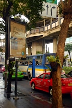 A busy urban street scene featuring a blue and yellow bus, red and green cars, and a large advertisement billboard. The advertisement is written in a foreign script. Elevated train tracks and traffic signs are visible above and around the vehicles. The scene is partially obscured by trees lining the street, and the road appears wet, suggesting recent rain.