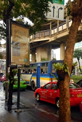 A busy urban street scene featuring a blue and yellow bus, red and green cars, and a large advertisement billboard. The advertisement is written in a foreign script. Elevated train tracks and traffic signs are visible above and around the vehicles. The scene is partially obscured by trees lining the street, and the road appears wet, suggesting recent rain.