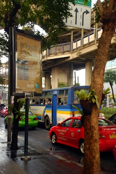 A busy urban street scene featuring a blue and yellow bus, red and green cars, and a large advertisement billboard. The advertisement is written in a foreign script. Elevated train tracks and traffic signs are visible above and around the vehicles. The scene is partially obscured by trees lining the street, and the road appears wet, suggesting recent rain.