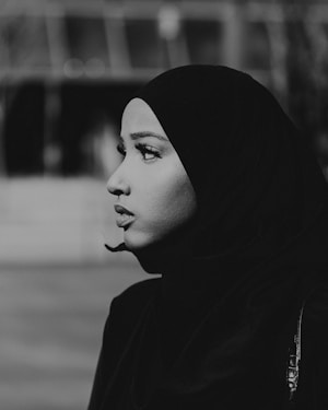 Profile view of a woman wearing a hijab, creating a strong contrast between the dark fabric and her facial features. The background is blurred, drawing attention to her contemplative expression.