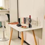 A modern white table with wooden legs is adorned with a variety of objects. There is a collection of colorful electric bottle openers, a bottle of red wine, two empty wine glasses, and a small plate with sliced cured meats. A cutting board with cherry tomatoes is placed on the table. In the background, a white bar stool is situated beside a cabinet with neatly arranged items and a plant.