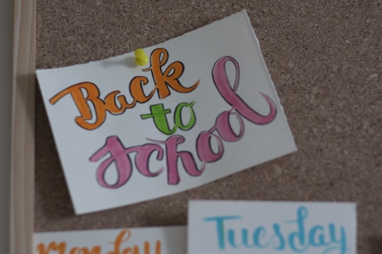 A corkboard features a piece of paper pinned with a yellow thumbtack. The paper has the words 'Back to school' written in colorful calligraphy, using orange, green, and pink shades. Below, partially visible days of the week are written in different colors. The setting conveys an organizational or planning theme.