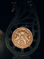 An ornate metalwork design with a golden circular emblem in the center, featuring a face. The intricate patterns in the metal include curves and flourishes, giving a decorative and antique appearance. The metal is dark with a weathered texture.