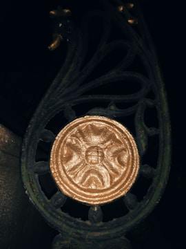 An ornate metalwork design with a golden circular emblem in the center, featuring a face. The intricate patterns in the metal include curves and flourishes, giving a decorative and antique appearance. The metal is dark with a weathered texture.