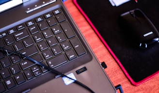 A close-up of a laptop keyboard in the foreground, with a numeric keypad featuring visible keys like 'Enter,' 'Delete,' and number pad digits. To the right, there is a black computer mouse placed on a black mouse pad with red trim. A laptop cable is also visible, crossing over the keyboard.