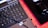 A close-up of a laptop keyboard in the foreground, with a numeric keypad featuring visible keys like 'Enter,' 'Delete,' and number pad digits. To the right, there is a black computer mouse placed on a black mouse pad with red trim. A laptop cable is also visible, crossing over the keyboard.