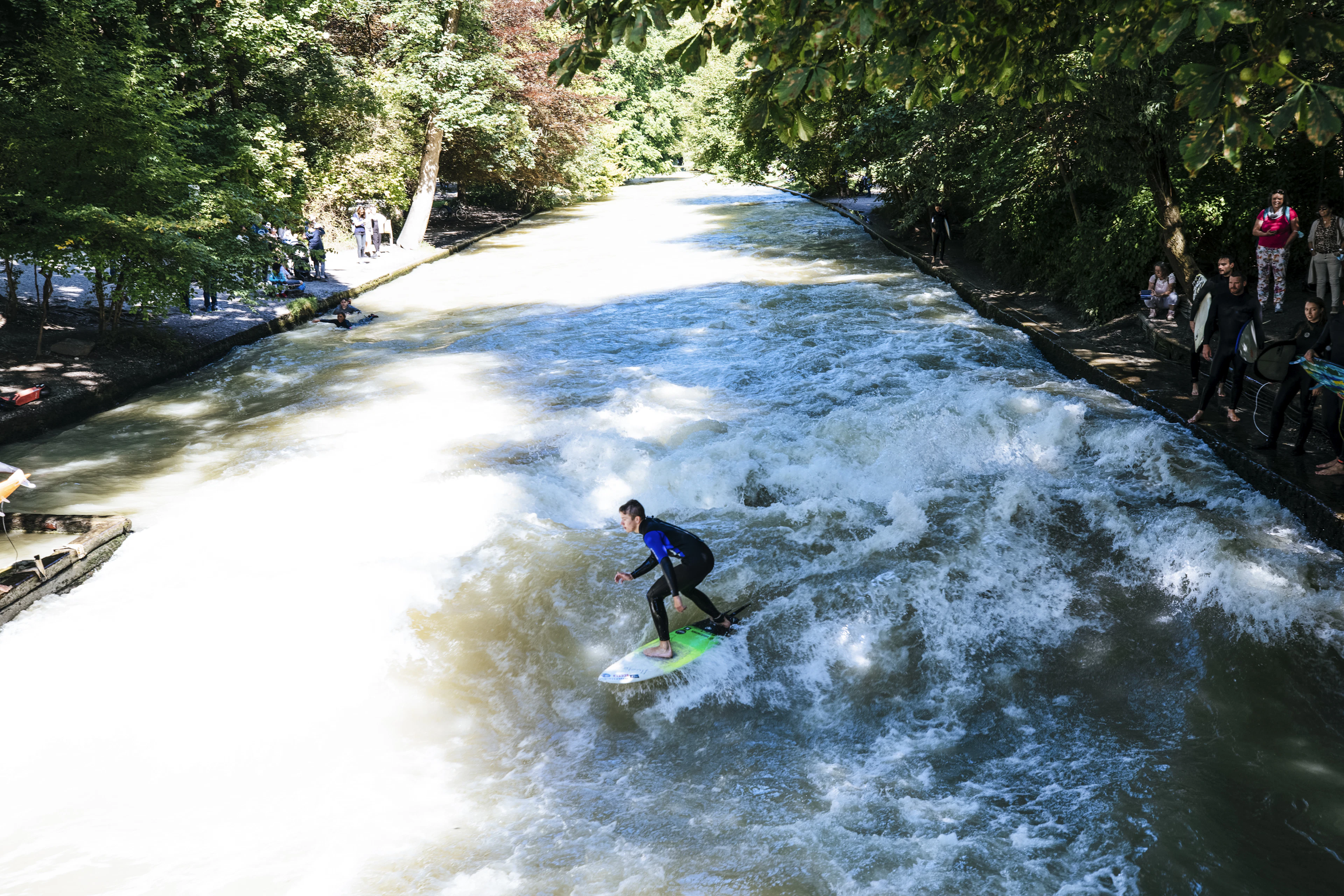 A wetsuited surfer riding the standing wave on the Eisbach in Munich's English Garden, white water churning down the narrow green channel while onlookers line the stone banks