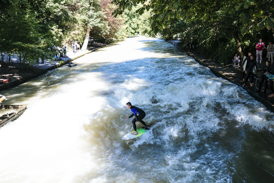 A surfer riding the standing wave on the Eisbach in Munich's English Garden, with onlookers on the stone banks