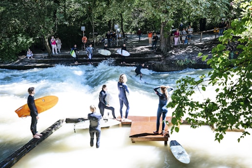 Surfers in wetsuits stand by a river with a strong current, while one surfer rides the waves. Spectators are gathered on the riverbank, observing the scene surrounded by lush green trees.