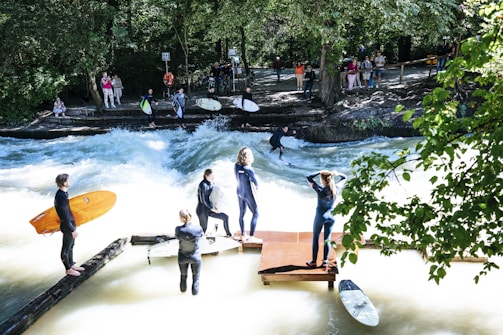 Surfers in wetsuits stand by a river with a strong current, while one surfer rides the waves. Spectators are gathered on the riverbank, observing the scene surrounded by lush green trees.