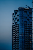 A modern high-rise building with sleek glass windows stands prominently against a dusky sky. The structure exhibits a mix of geometric shapes and reflective surfaces with a construction crane visible on top, suggesting ongoing development.