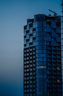 A modern high-rise building with sleek glass windows stands prominently against a dusky sky. The structure exhibits a mix of geometric shapes and reflective surfaces with a construction crane visible on top, suggesting ongoing development.