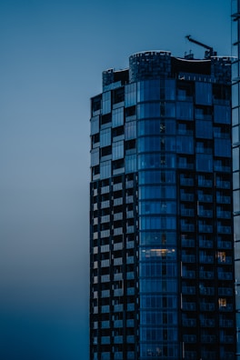 A modern high-rise building with sleek glass windows stands prominently against a dusky sky. The structure exhibits a mix of geometric shapes and reflective surfaces with a construction crane visible on top, suggesting ongoing development.
