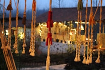 A cozy outdoor setting with hanging rope decorations featuring tassels in various colors like red and natural tones. In the background, a warmly lit patio or porch is visible with string lights and rustic furniture, creating an inviting atmosphere.