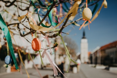 Colorful Easter eggs and ribbons are hanging from tree branches, with a blurred background featuring a tall building with a spire and an out-of-focus street scene.