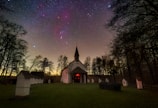 A church with a steeple is nestled in a wooded area under a clear night sky filled with stars. The sky is vibrant with scattered stars against a faintly visible Milky Way. The foreground features a grassy area leading up to the church, surrounded by silhouetted trees and small white shrines or stations.