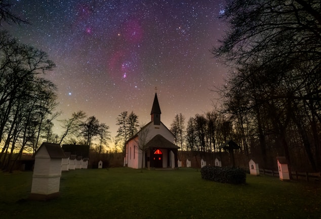A church with a steeple is nestled in a wooded area under a clear night sky filled with stars. The sky is vibrant with scattered stars against a faintly visible Milky Way. The foreground features a grassy area leading up to the church, surrounded by silhouetted trees and small white shrines or stations.
