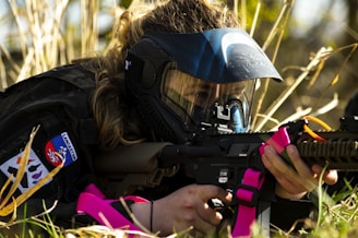 A person wearing military-style attire and a protective mask is lying on the ground amidst tall grass while aiming a rifle. The rifle is adorned with bright pink elements, possibly straps. The attire includes patches, one of which has some text and symbols.