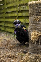 A person wearing a tactical outfit, including a helmet with a camera and a face mask, is crouching behind a haystack in an outdoor area. The person is holding a paintball gun and equipped with gear suitable for an action-packed activity. The scene is set against a background of a wooden fence and a ground covered with dead grass and foliage.