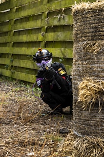A person wearing a tactical outfit, including a helmet with a camera and a face mask, is crouching behind a haystack in an outdoor area. The person is holding a paintball gun and equipped with gear suitable for an action-packed activity. The scene is set against a background of a wooden fence and a ground covered with dead grass and foliage.