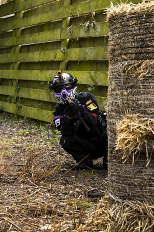 A person wearing a tactical outfit, including a helmet with a camera and a face mask, is crouching behind a haystack in an outdoor area. The person is holding a paintball gun and equipped with gear suitable for an action-packed activity. The scene is set against a background of a wooden fence and a ground covered with dead grass and foliage.