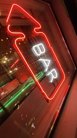A neon sign with the word 'BAR' illuminated in white letters outlined by a red arrow. The sign is displayed against a glass window, reflecting the lights. There are additional green and orange lights creating an urban nightlife ambiance.