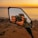 A camera is held up by a person capturing a reflection in a side mirror. The background features a scenic view of a body of water and a vibrant sunset sky. The scene is calm and picturesque, conveying a moment of photography during a sunset.